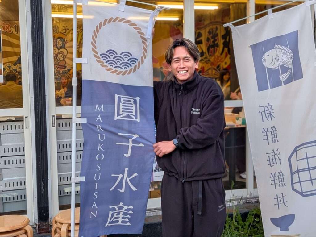 「圓子水産」店舗前でのぼりを持ちながら笑う圓子瑞樹さん